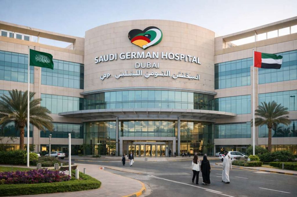 Exterior view of Saudi German Hospital Dubai showing the main entrance, hospital logo, glass facade, landscaped garden, flags, and people walking towards the hospital